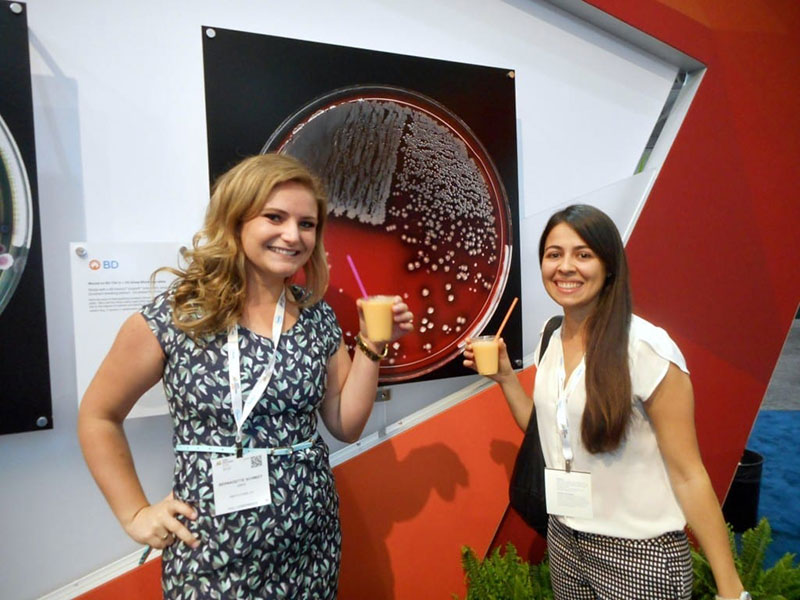 BREAK TIME: UMHS students Bernadette Schmidt & Victoria Gonzalez at the BD Diagnostics (Becton, Dickinson and Company) booth at the conference vendor fair. 'BD is the manufacturer of the blood agar plates we used in our experiments to gain results similar to the hemolytic Staph aureus as pictured on the wall print,' says Dr. Harrington. Photo: Dr. Jane Harrington
